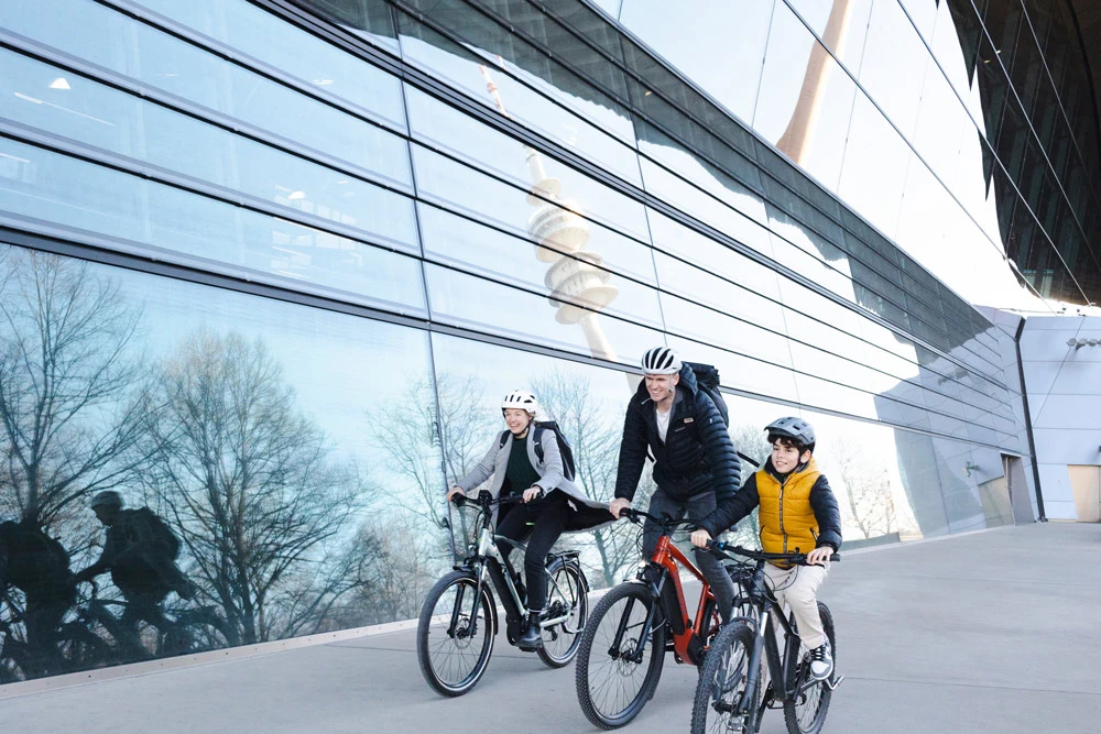 Eine dreiköpfige Familie fährt auf Fahrrädern vor einem modernen Glasgebäude, in dem sich Bäume und Fahrräder spiegeln.