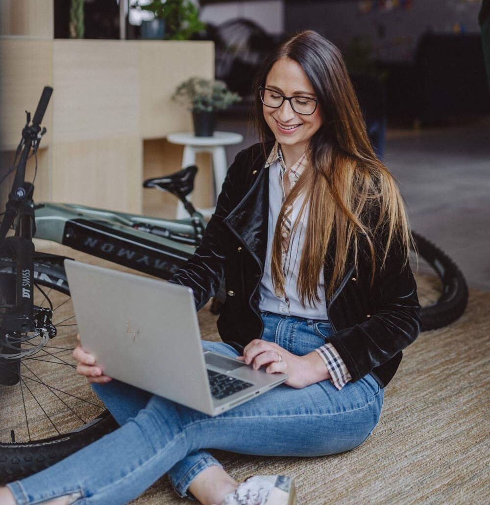 Eine Frau sitzt auf dem Boden neben einem Fahrrad und arbeitet lächelnd an ihrem Laptop. Im Hintergrund sind Pflanzen und ein moderner, offener Raum zu sehen.