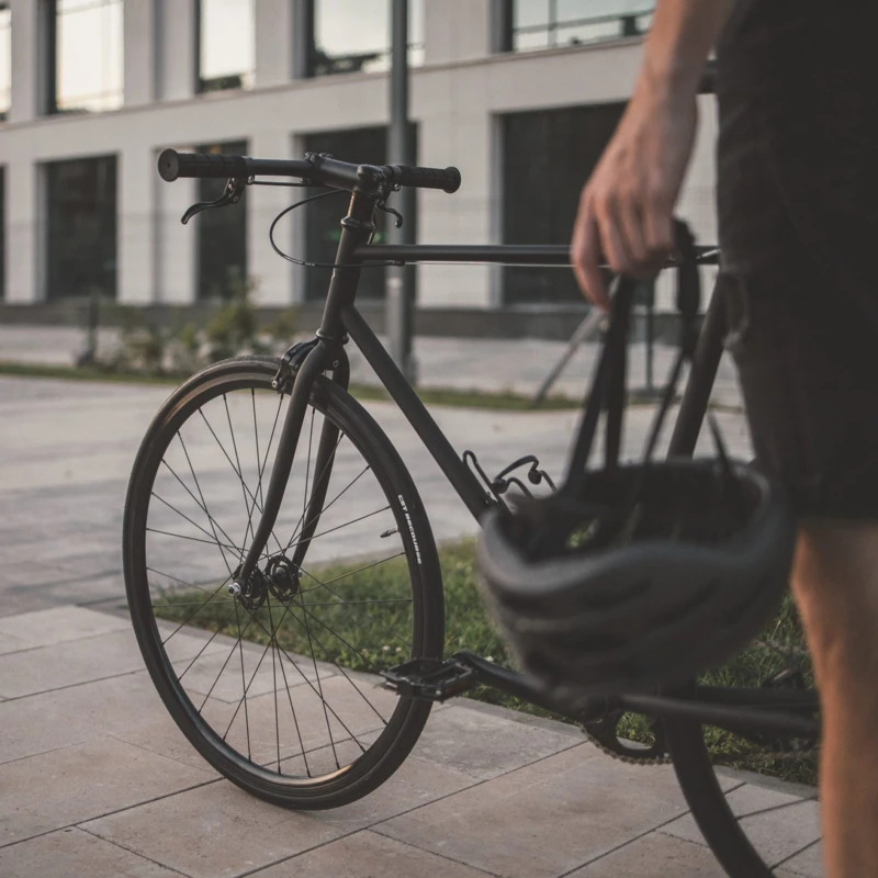 Eine Person hält einen Fahrradhelm und steht neben einem schlanken, schwarzen Fahrrad in einem modernen städtischen Umfeld.